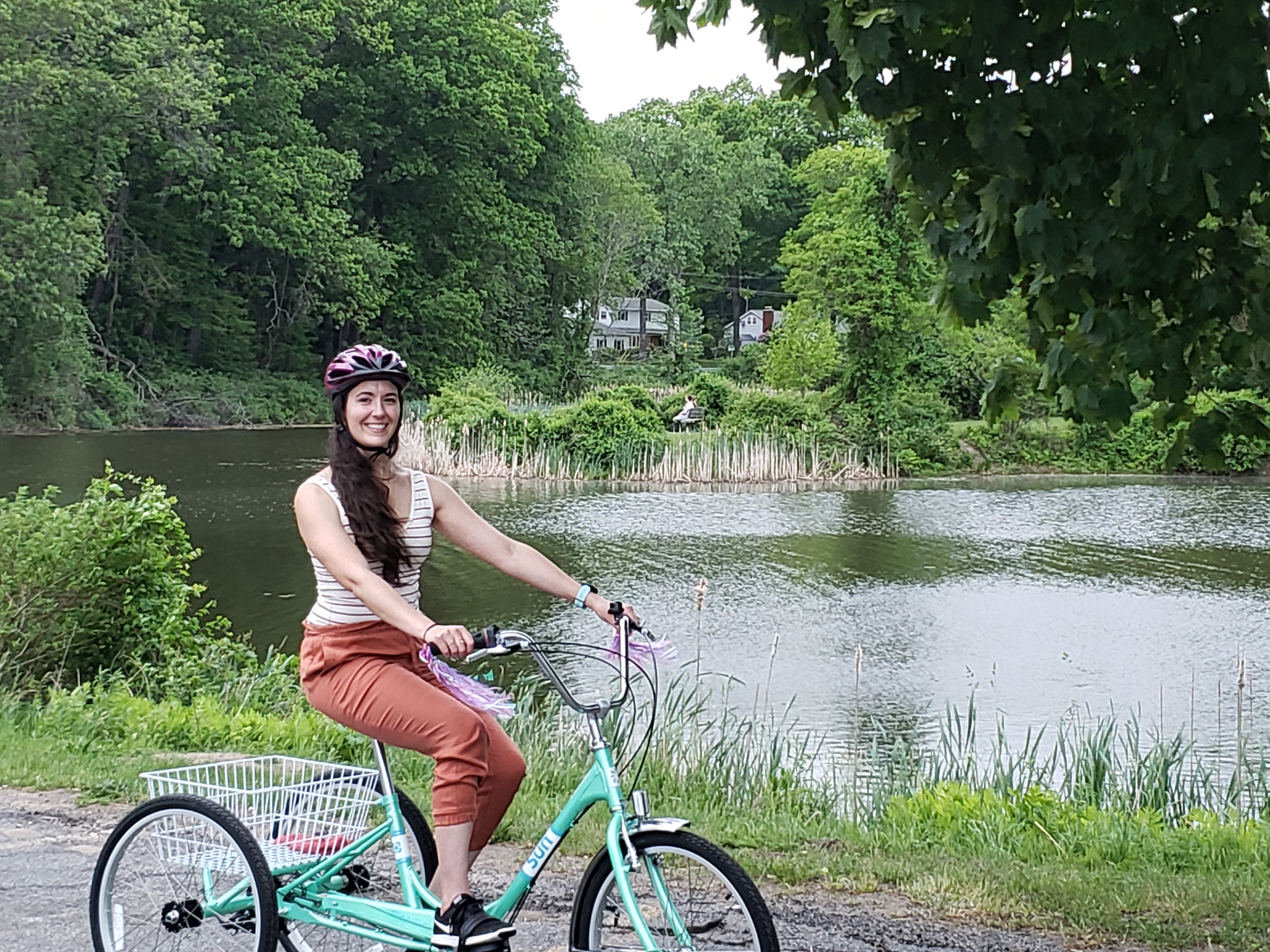 Kerry in copper colored pants and a white and red striped tank top, and a purple helmet, on her mint green tricycle, in front of a lake/pond surrounded by green trees.